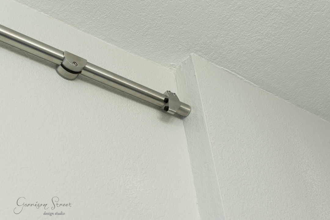 Close-up of a steel curtain rod mounted in the corner of a white ceiling, reminiscent of sleek barn door hardware. The rod is secured with two brackets, and the wall has a slight texture. A logo for Garrison Street Design Studio is visible in the bottom left corner.