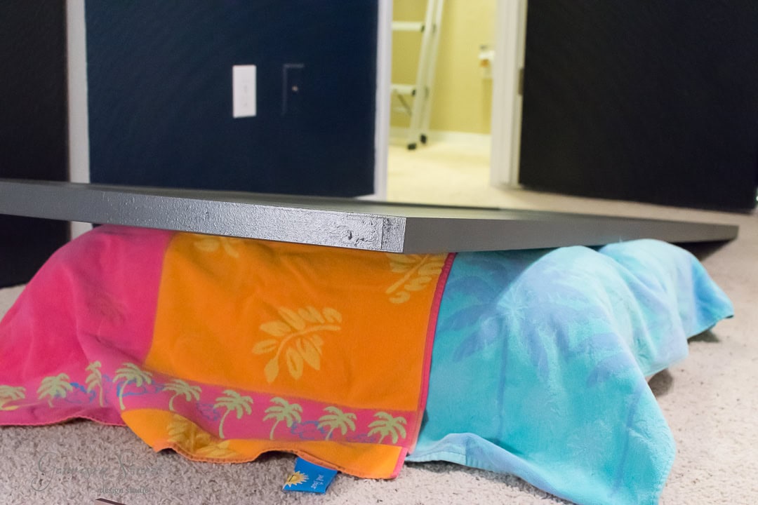 A pastel-colored beach towel, featuring orange, pink, and blue sections with palm tree patterns, props up a large, flat piece of wood reminiscent of a barn door on a carpeted floor. An open doorway and part of a ladder are visible in the background.