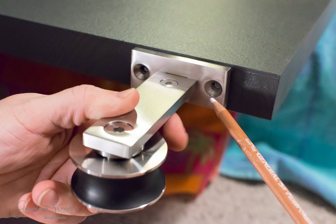 Close-up of a hand holding a barn door roller assembly against the edge of a black tabletop. A pencil is pointing at the screw hole on the metal bracket for precise alignment. The background is blurred with colorful elements.