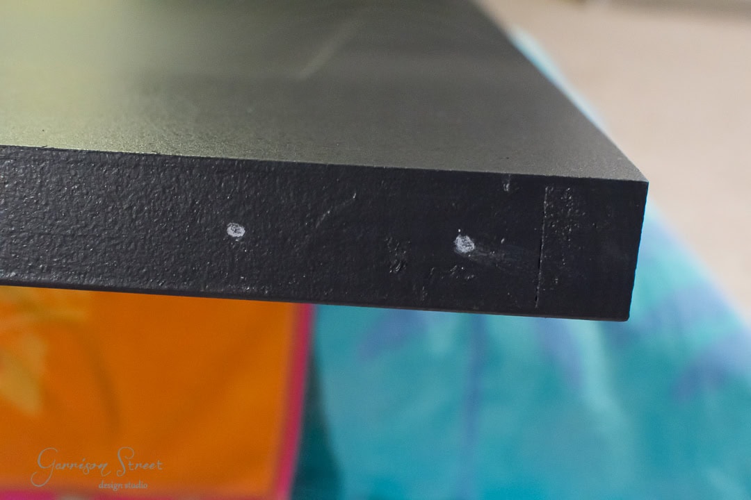 Close-up of the edge of a black tabletop with two small white markings on it, resembling the subtle charm of a rustic barn door. The background is blurred and colorful, featuring shades of blue and orange.