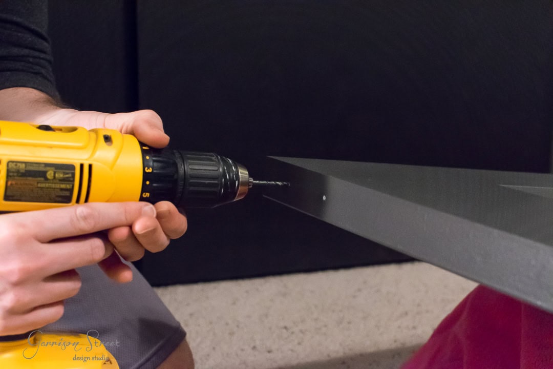 A person uses a yellow drill to make a hole in a dark-colored barn door, stabilizing the surface with their other hand. The focus is on the drill, held horizontally.