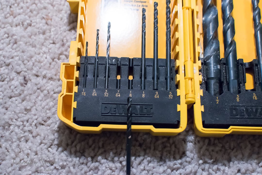 A yellow DeWalt drill bit set is open on a carpeted floor, displaying various sized drill bits like the organized planks of a barn door, each resting in its respective slot marked by size numbers.