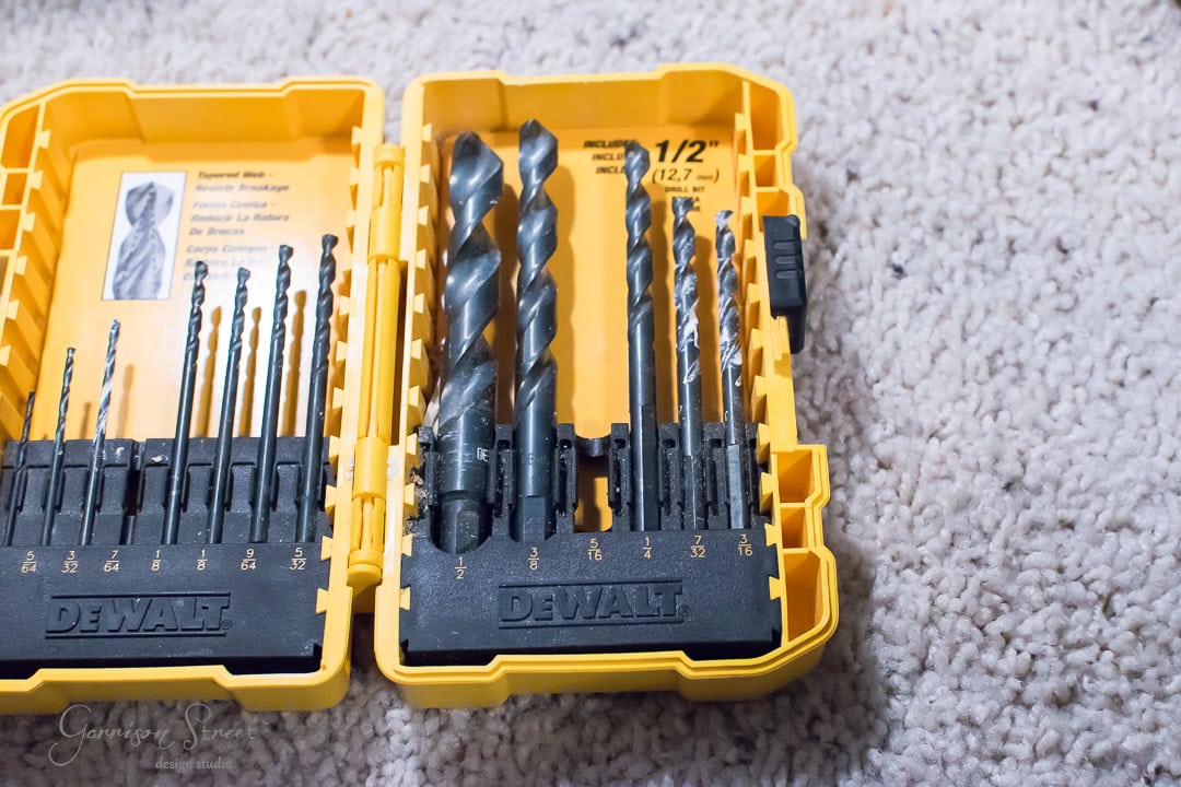 A yellow DeWalt drill bit set with various sizes is neatly arranged in a plastic case, resting on a light-colored carpet. The case's cover reveals labeled drill sizes and types, making it perfect for anyone ready to tackle projects like installing a rustic barn door.