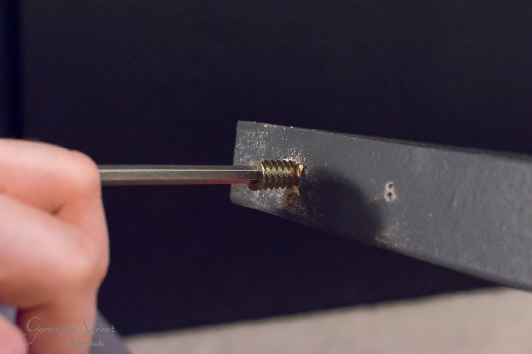A close-up of a hand holding a screwdriver, working on a screw set into a dark metal barn door surface. The focus is on the screw and screwdriver tip, with the rustic texture of the door softly blurred in the background.