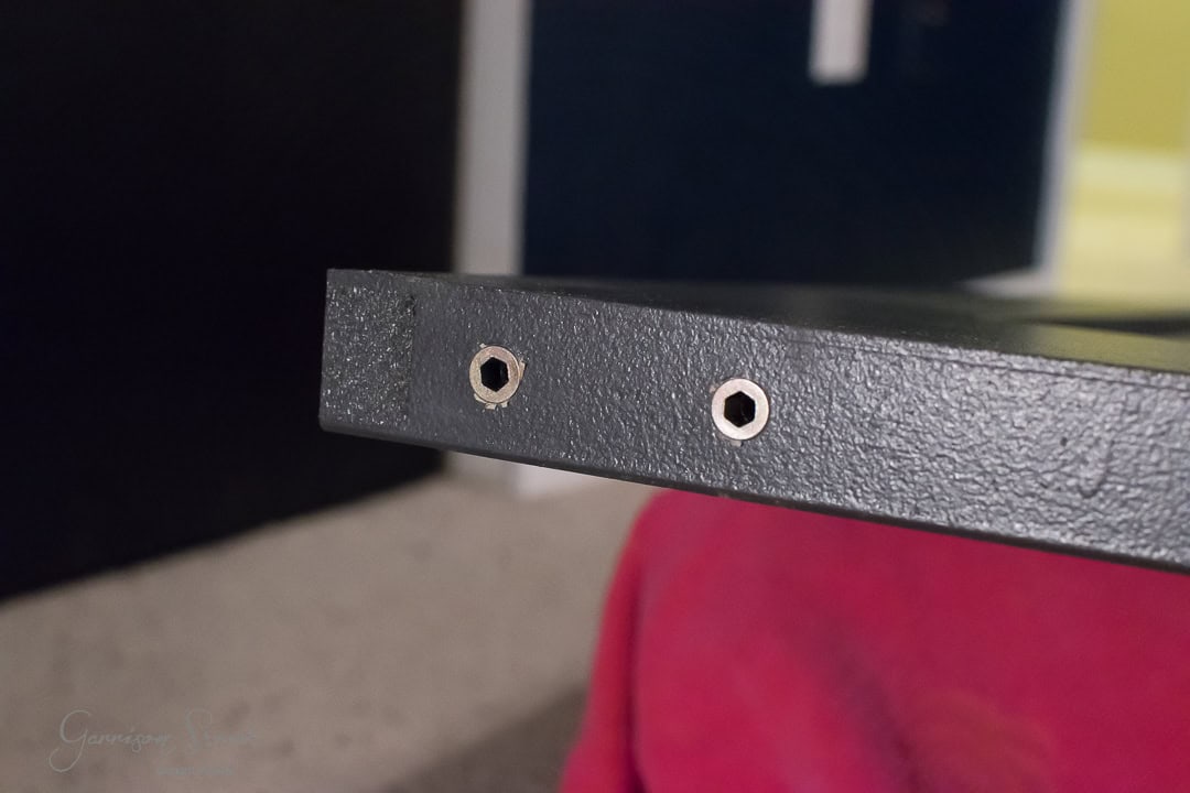 Close-up of the end of a black wooden barn door with two metal screw holes. The board stands vertically on a red fabric surface, against a blurry background revealing parts of a room.