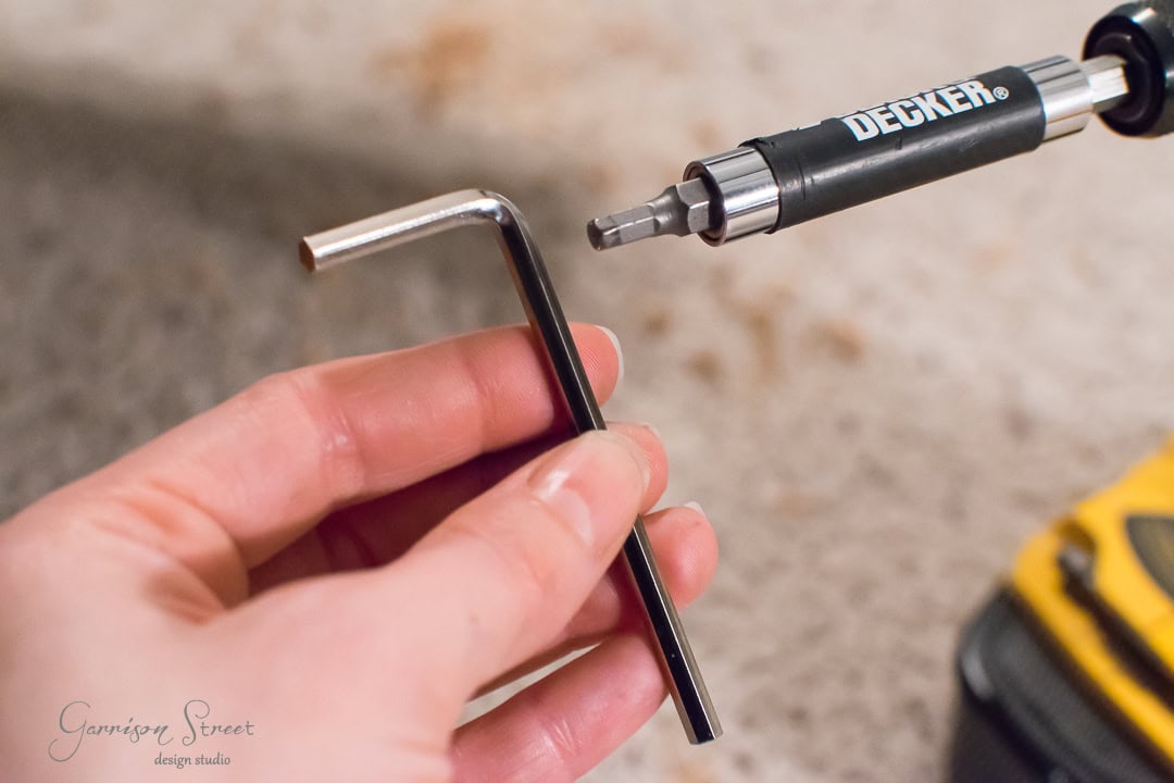 A hand grips an L-shaped hex key, while a cordless screwdriver with a hex bit rests nearby, ready to tackle the barn door assembly. A tool case is partially visible in the background.
