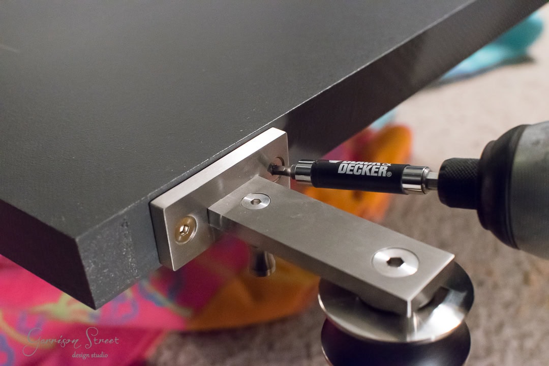 A close-up of a power drill with a Black & Decker bit attached, driving a screw into a metal bracket on the edge of a dark wood panel, reminiscent of barn door construction. A colorful fabric is slightly visible in the background.