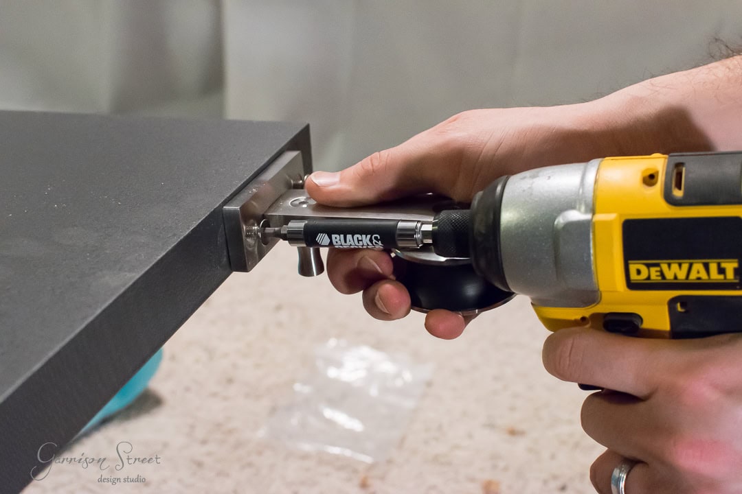 A person uses a yellow DeWalt drill to assemble a piece of furniture, effortlessly attaching a hinge to a dark-colored board, reminiscent of barn door wood. The word "BLACKS" is visible on the tool, while a countertop and small plastic bag sit subtly in the background.