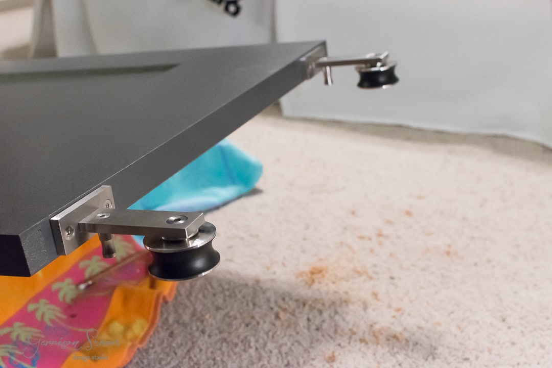 Close-up of a black tabletop with metal brackets and black wheels attached to its edge, reminiscent of a barn door. The background reveals a carpeted floor adorned with colorful fabric and a blue plastic bag.