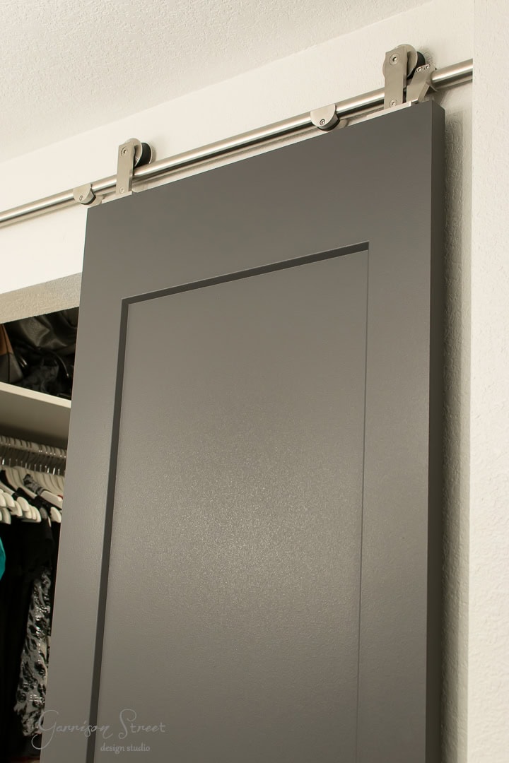 A sleek, dark gray barn door on a sliding metal track is partially open, revealing a closet with clothes hanging inside. The modern design beautifully contrasts against the crisp white walls.