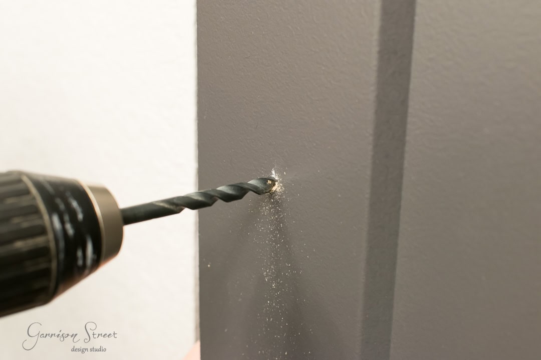 Close-up of a drill making a hole in a gray wall, with dust particles visible around the bit. The drill is angled as if crafting an entryway for a barn door, blending utility with rustic charm.