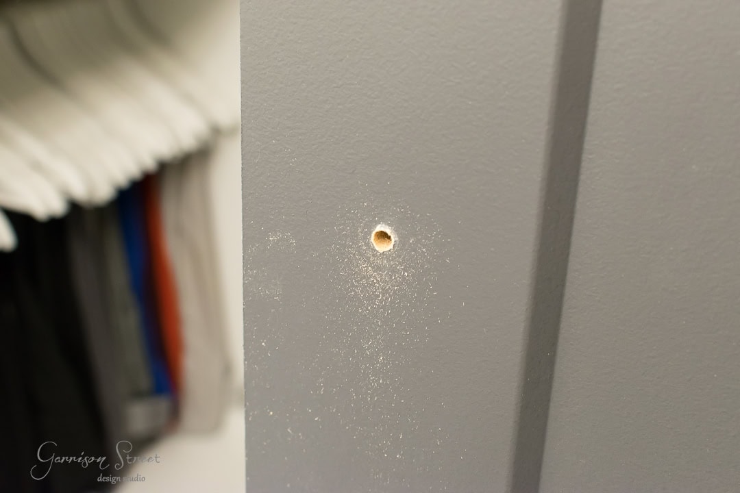 A close-up of a gray wall with a small circular hole drilled into it, reminiscent of the rustic charm of a barn door. Fine wood dust is scattered around the hole, while blurred in the background are some clothes hanging in a closet.