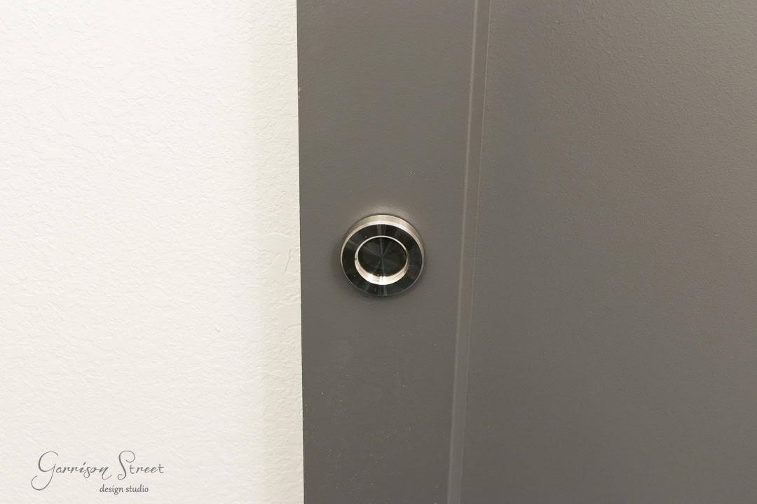 A sleek, modern silver doorbell is installed on a gray wall, reminiscent of a contemporary barn door aesthetic. The background features a subtle white texture on the left side, with "Garrison Street Design Studio" in small text below.