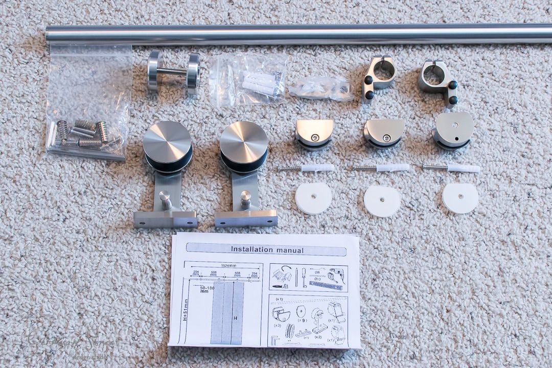A barn door hardware kit is elegantly displayed on a carpet. It features metal brackets, rollers, screws, spacers, and an installation manual, ensuring easy assembly. At the top of the image lies a long track essential for your barn door setup.