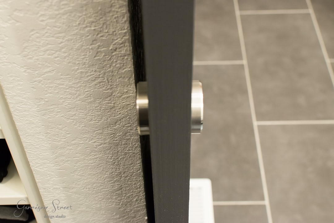 Close-up of a modern barn door with a metallic handle, partially open, revealing a textured wall to the left and gray tiled flooring in the background.