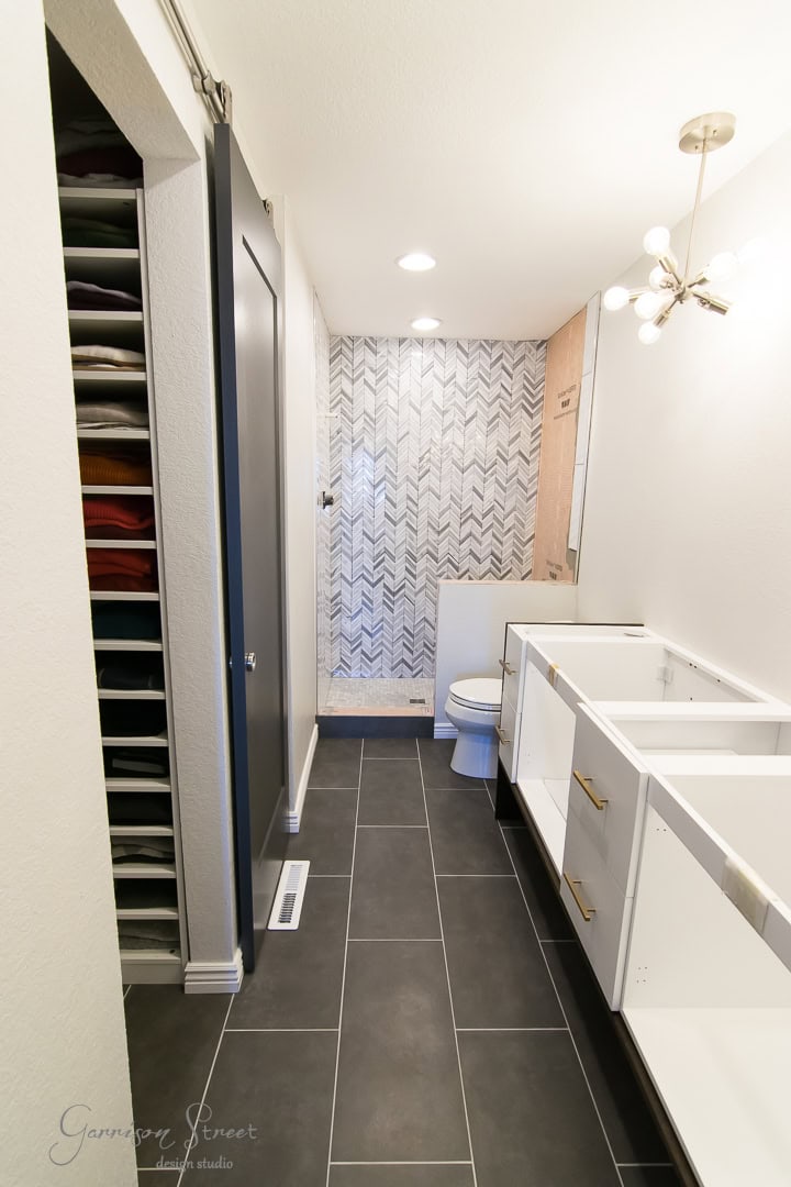 A modern bathroom under renovation, now featuring an elegant barn door. It boasts an empty vanity with open cabinets and gold hardware, a tiled herringbone shower with a glass door, black floor tiles, and wall-mounted light fixtures. Towels are neatly stacked on an open shelf..
