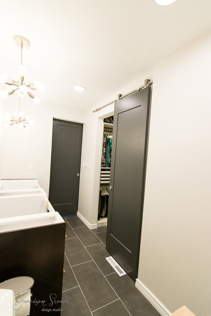 The modern bathroom showcases a sleek gray tile floor and pristine white walls. A striking dark barn door slides open to reveal a closet filled with neatly hung clothes, while a white vanity and unique light fixture add sophistication to the space.