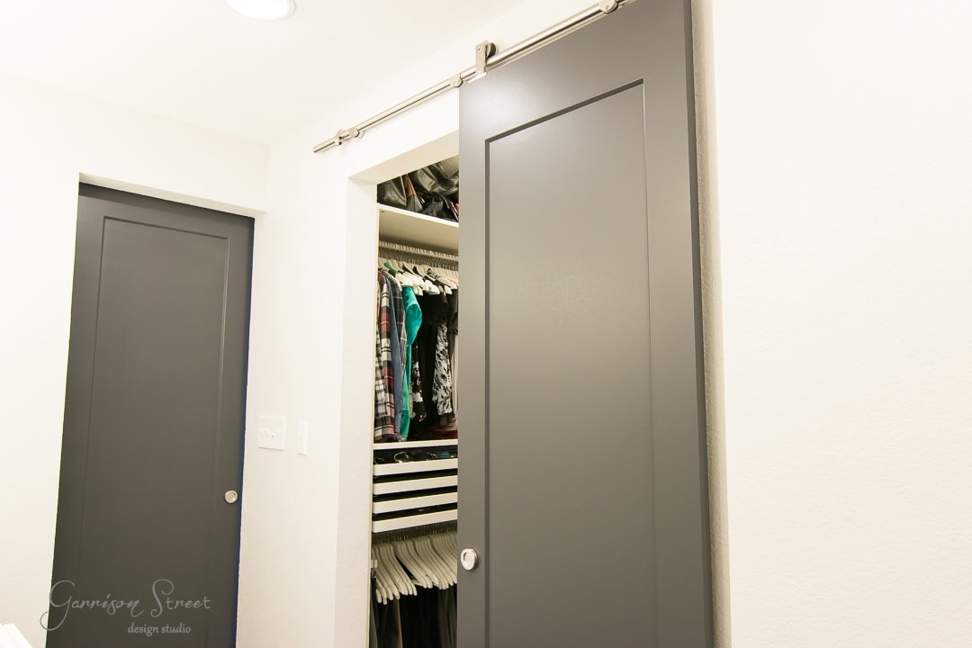 A modern closet with sliding gray barn doors open to reveal a neatly organized interior. Clothes hang on rods, with shirts, pants, and other garments visible. The walls and ceiling are white, giving a clean and minimalist look.