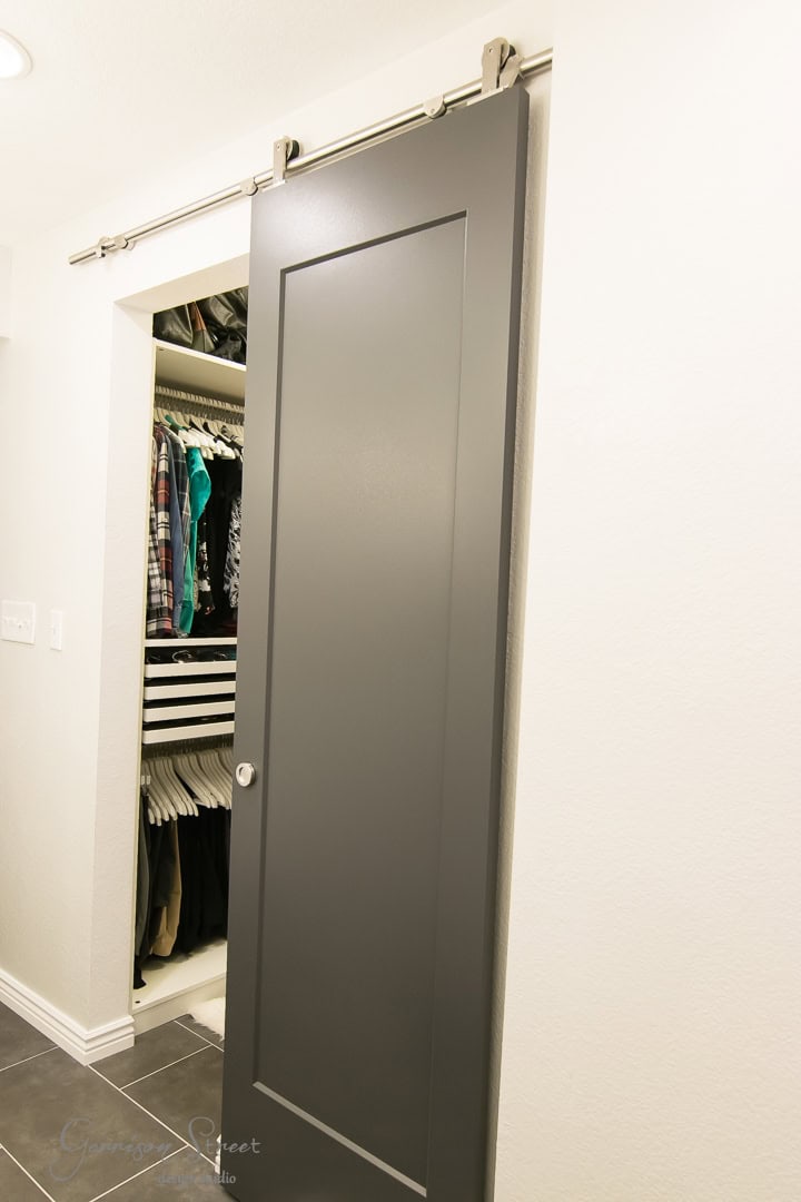 A sleek gray barn door, partially open, reveals a neatly organized closet with clothes hanging and storage shelves. The modern metal hardware of the barn door adds a contemporary touch, while the dark-tiled floor enhances the room's elegance.