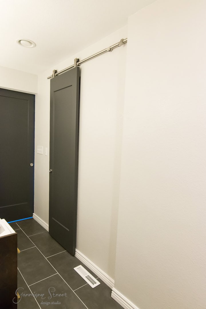 A minimalist hallway showcases a sleek barn door sliding smoothly on a metal track against a light gray wall. With dark gray tiles underfoot and a crisp white baseboard, the space merges rustic charm with modern elegance.