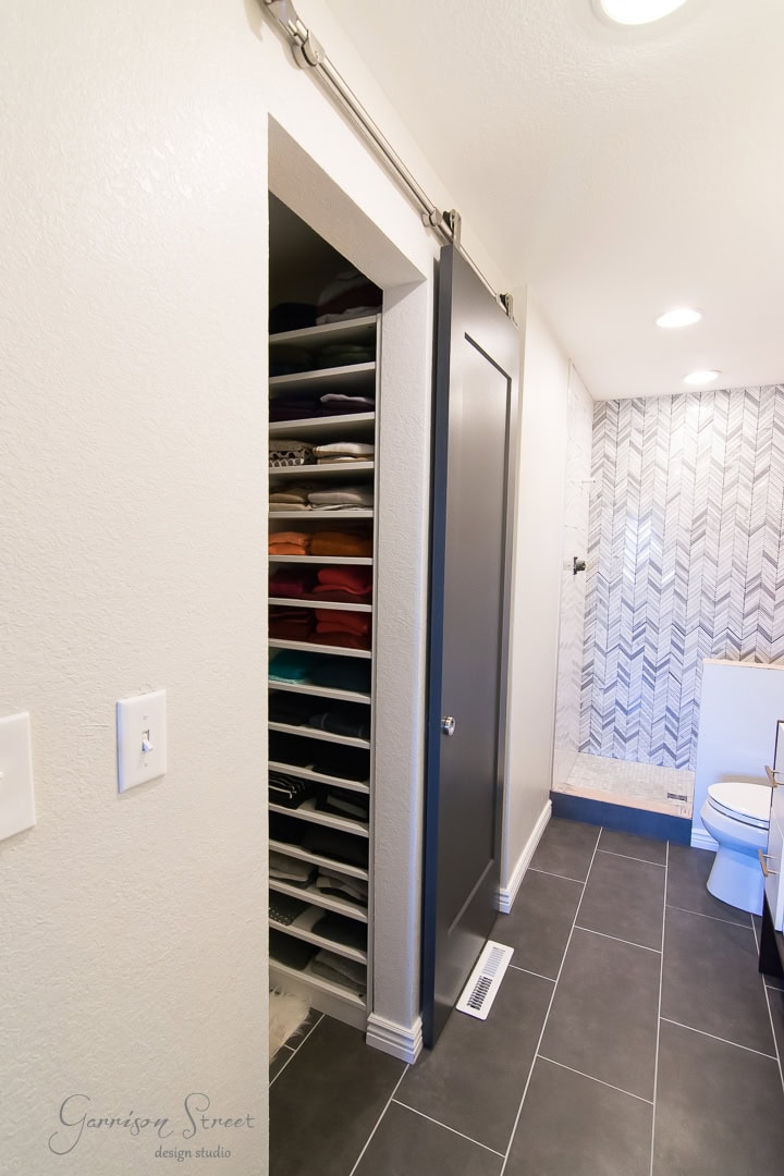 A modern bathroom with dark floor tiles and a sliding barn door reveals a neatly organized closet filled with colorful towels. The wall features a herringbone tile pattern, complementing the stylish design, and a toilet is subtly tucked in the corner.