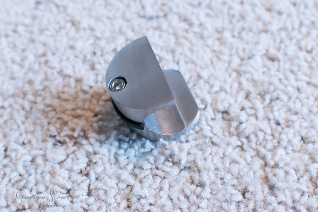 A small, metallic object with a rounded edge and a screw visible on top, resting like a forgotten treasure on a textured, barn door-hued carpet.