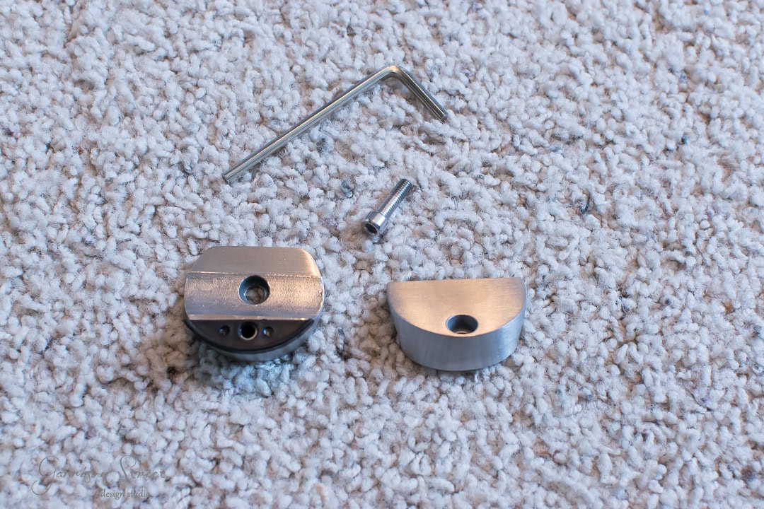 Two metallic components and a hex key rest on a textured carpet, reminiscent of pieces from a barn door assembly. The components have multiple holes for assembly, with a small cylindrical metal piece visible between them.