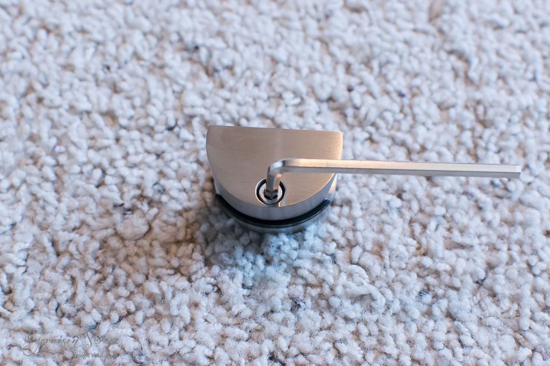 Close-up of a small metal tool with an L-shaped hex key inserted into a round, brushed metal base, reminiscent of mechanisms found in barn door hardware. The object rests on a textured, light-colored carpet.