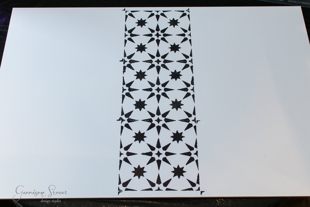 A white canvas with a vertical strip of black stencil design featuring repeating stars and geometric patterns in the center.