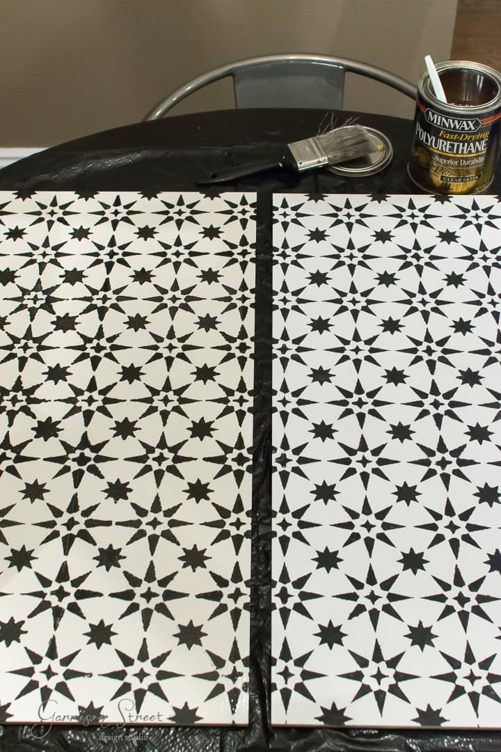 A table is covered with stenciled tiles featuring a black starburst pattern on a white background. A can of Minwax polyurethane and a paintbrush are placed on the table. A metal chair is partially visible.