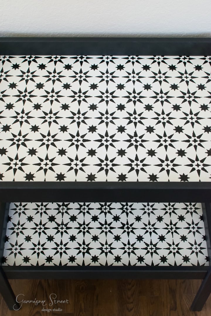 A table with a black frame features a white surface decorated with a geometric star pattern in black. The lower shelf matches the design. The floor is wooden.
