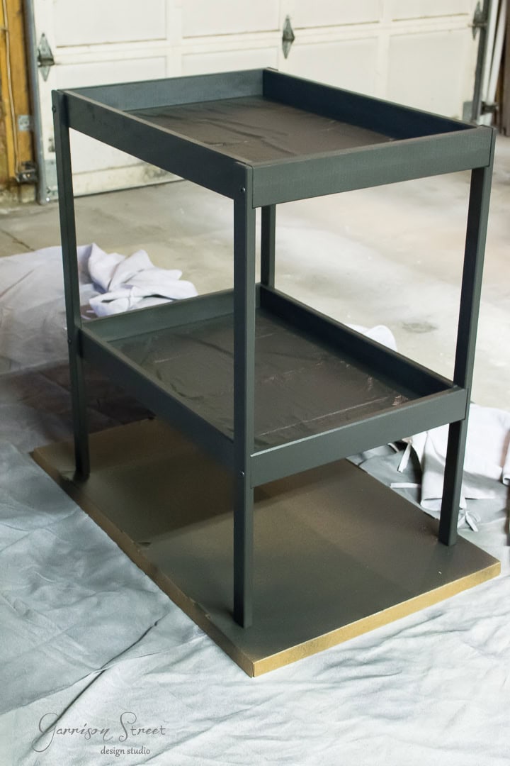 A small, dark green, two-tier metal cart with a wooden base sits in a garage. The garage has a concrete floor, and there's a cloth spread on the ground.