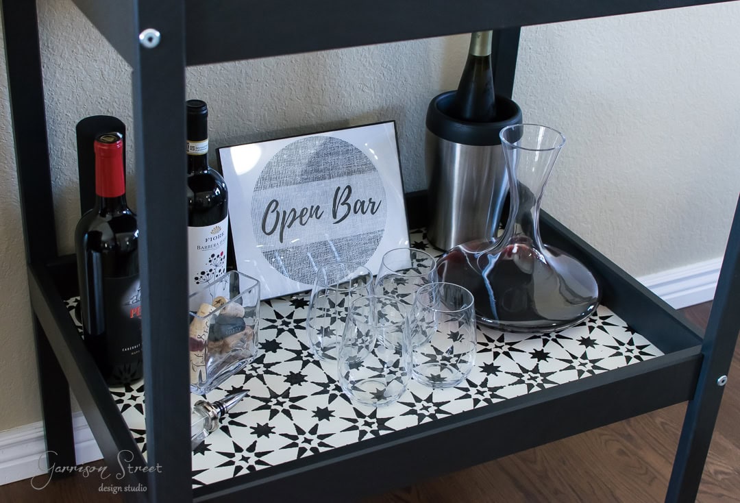 A bar cart with wine bottles, a decanter, glassware, and a small sign that reads "Open Bar" on a patterned shelf.