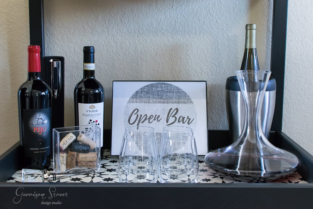 A bar setup with two wine bottles, a wine decanter, two empty glasses, an ice bucket, and a sign reading "Open Bar." There are also wine corks and a small decorative box on a patterned tray.