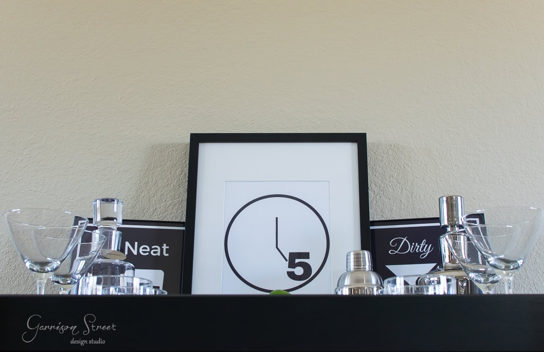A bar shelf displays cocktail shakers, martini glasses, and a framed minimalist clock print showing 5 o'clock. Signs labeled "Neat" and "Dirty" are on either side of the print. A lime is placed in front of the frame.