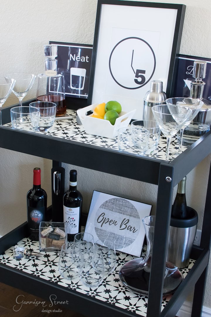 A stylish bar cart features glasses, bottles of liquor, a wine bottle, a shaker, and a framed clock print. There's a bowl of lemons and limes, along with a sign that says "Open Bar". The surface has a geometric black and white pattern.