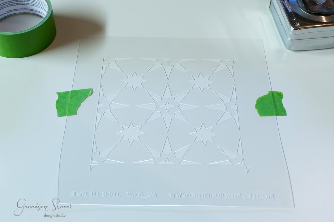 A square, transparent stencil with star patterns taped to a white surface using green tape. A roll of green tape and part of a metal object are visible nearby.