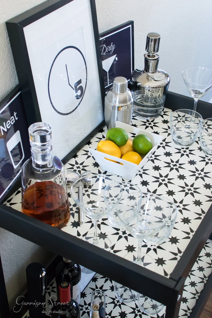 A stylish black bar cart features a glass decanter, cocktail shaker, martini glasses, and a bowl of lemons and limes. Decorative signs saying "Neat" and "Dirty" are displayed, with a framed image showing a clock set at 5 o'clock.