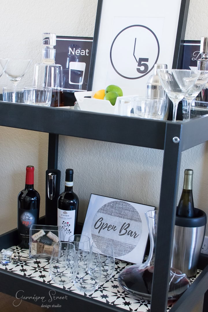 A stylish bar cart with various glasses, bottles of wine, a shaker, and a wine cooler. Decorative signs and a clock are displayed on top. Lemons and limes add a pop of color. The lower shelf features an "Open Bar" sign.