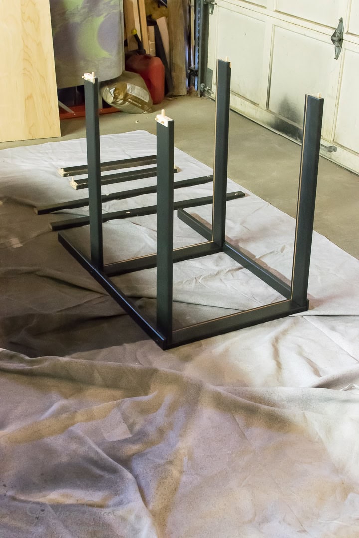 A partially assembled black metal table frame lies upside down on a tarp in a garage. Surrounding the frame are disassembled wooden panels and tools, suggesting ongoing construction or assembly.