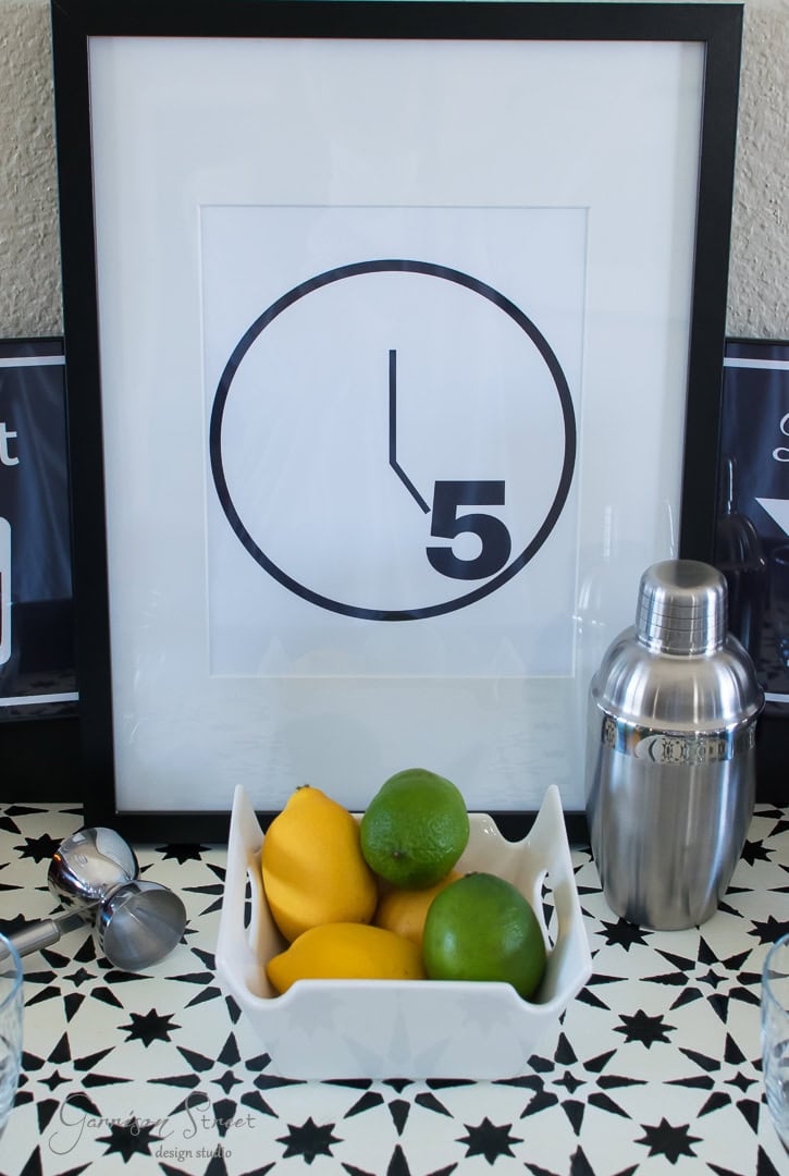 A framed print with a clock design and the number "5" is on a patterned table. In front, there's a bowl with lemons and limes, a cocktail shaker, sunglasses, and two glasses.