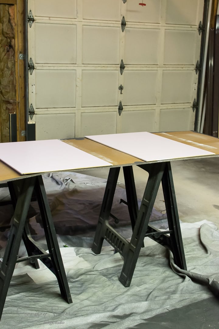 A garage workspace with two sawhorses holding a large piece of plywood, covered partially with three white boards. A drop cloth is spread on the floor underneath, suggesting preparation for painting or a project.