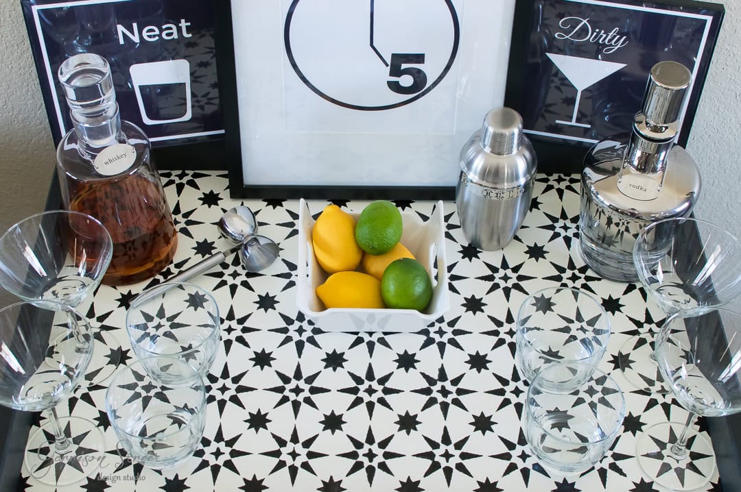 A bar setup with whiskey and vodka bottles, a cocktail shaker, lemons, and a lime on a patterned black and white table. Four glasses are arranged around the table. Wall signs read "Neat" and "Dirty" with cocktail illustrations.