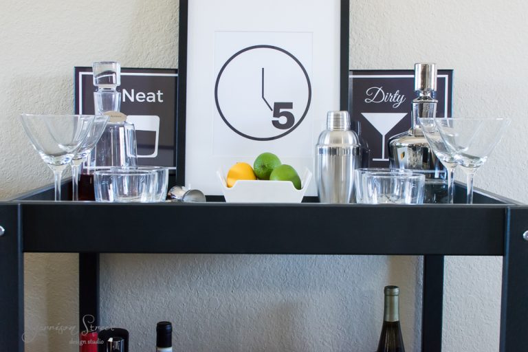 A bar cart with various cocktail glasses, shakers, bottles of liquor, and a bowl of lemons and limes. Behind the cart are two framed signs with cocktail illustrations and a clock showing 5 o'clock.