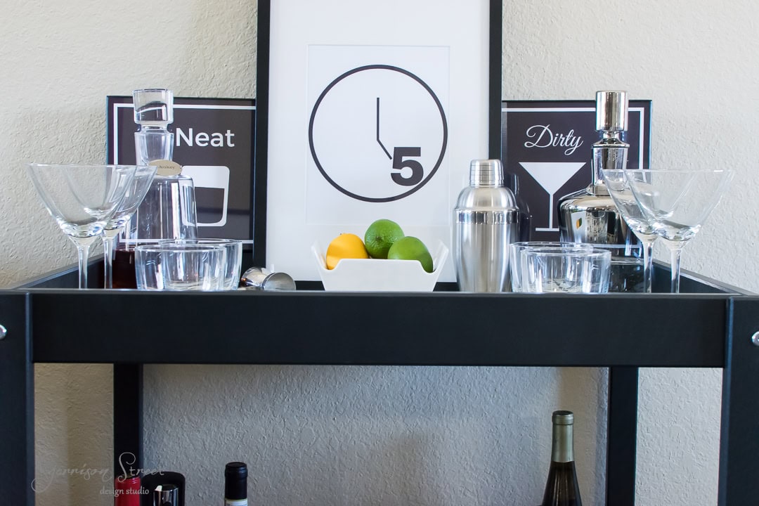 A bar cart with various cocktail glasses, shakers, bottles of liquor, and a bowl of lemons and limes. Behind the cart are two framed signs with cocktail illustrations and a clock showing 5 o'clock.