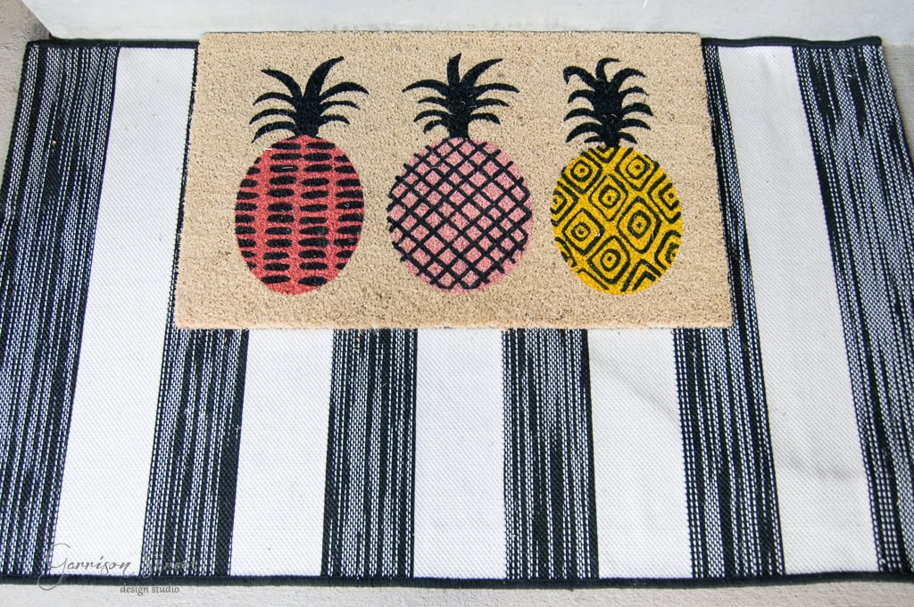 Doormat featuring three pineapples in red, pink, and yellow with unique patterns on each, set against a black and white striped background.