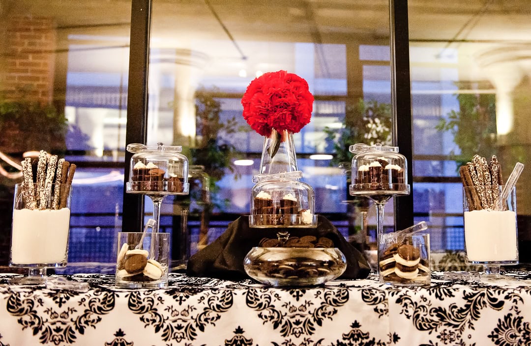 A dessert table with a black and white patterned cloth featuring cookies, cupcakes, and chocolate-dipped pretzels in glass jars. A vase with red flowers is centered at the back. The background shows a window and plants.