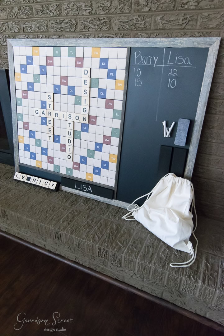 A large Scrabble board mounted on a black board with scores tallying for Barry and Lisa on the side. Below, a white drawstring bag and Scrabble tiles spelling "LUCKY" and "LISA" are placed. The setup rests against a brick wall.
