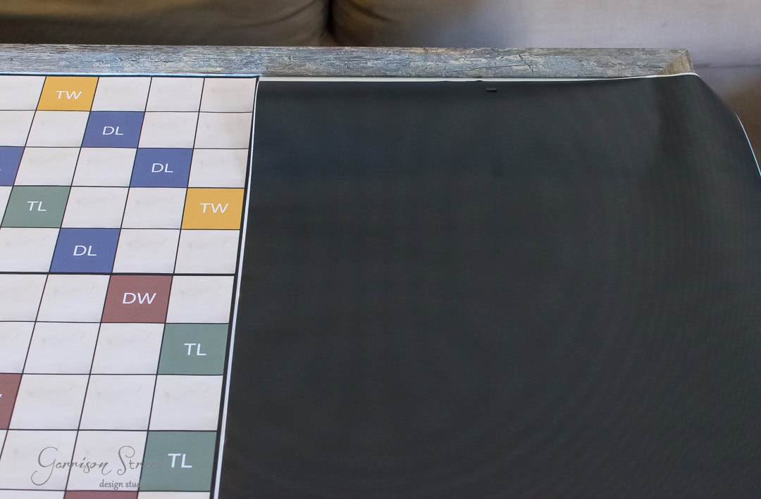 Close-up of a partially unrolled Scrabble game board on a table. The board displays various letter score squares, such as "TW" for triple word and "DL" for double letter. There's an empty space on the right side of the board.