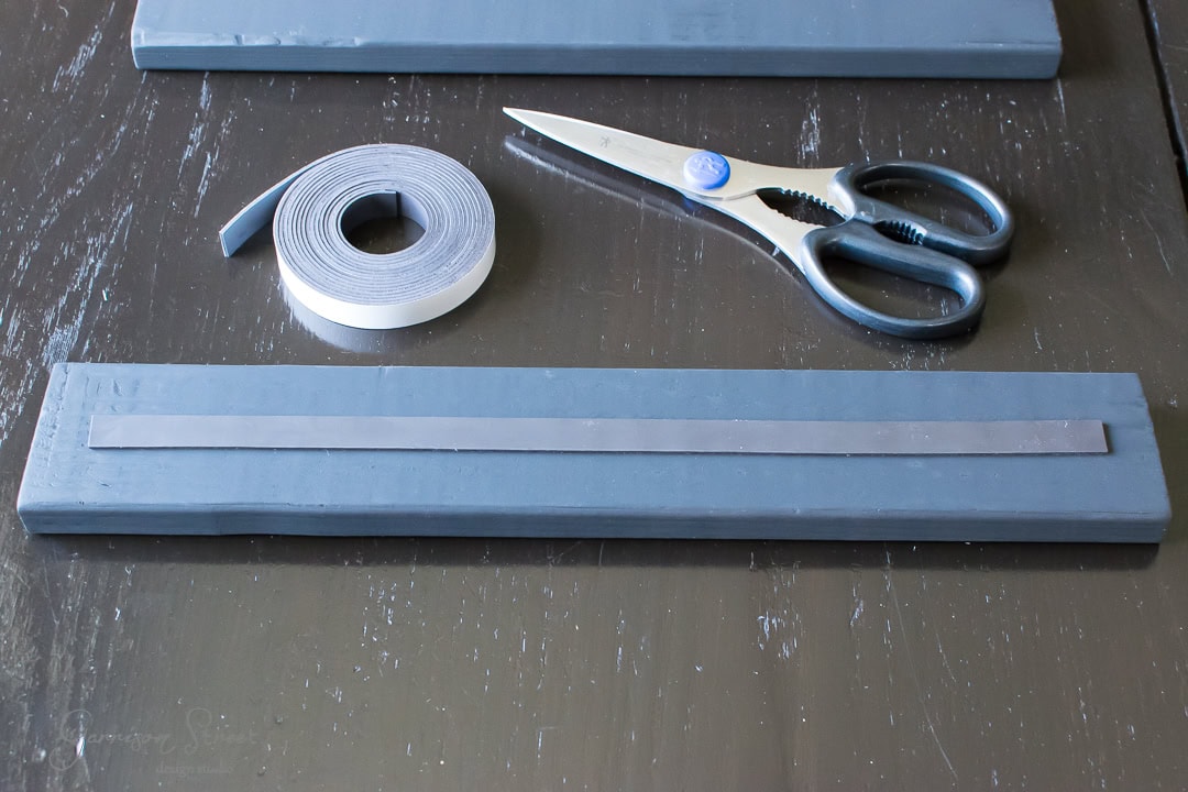 A pair of scissors, a roll of adhesive magnetic strip, and two rectangular magnetic strips laid on a dark wooden surface. One strip is partially unrolled.